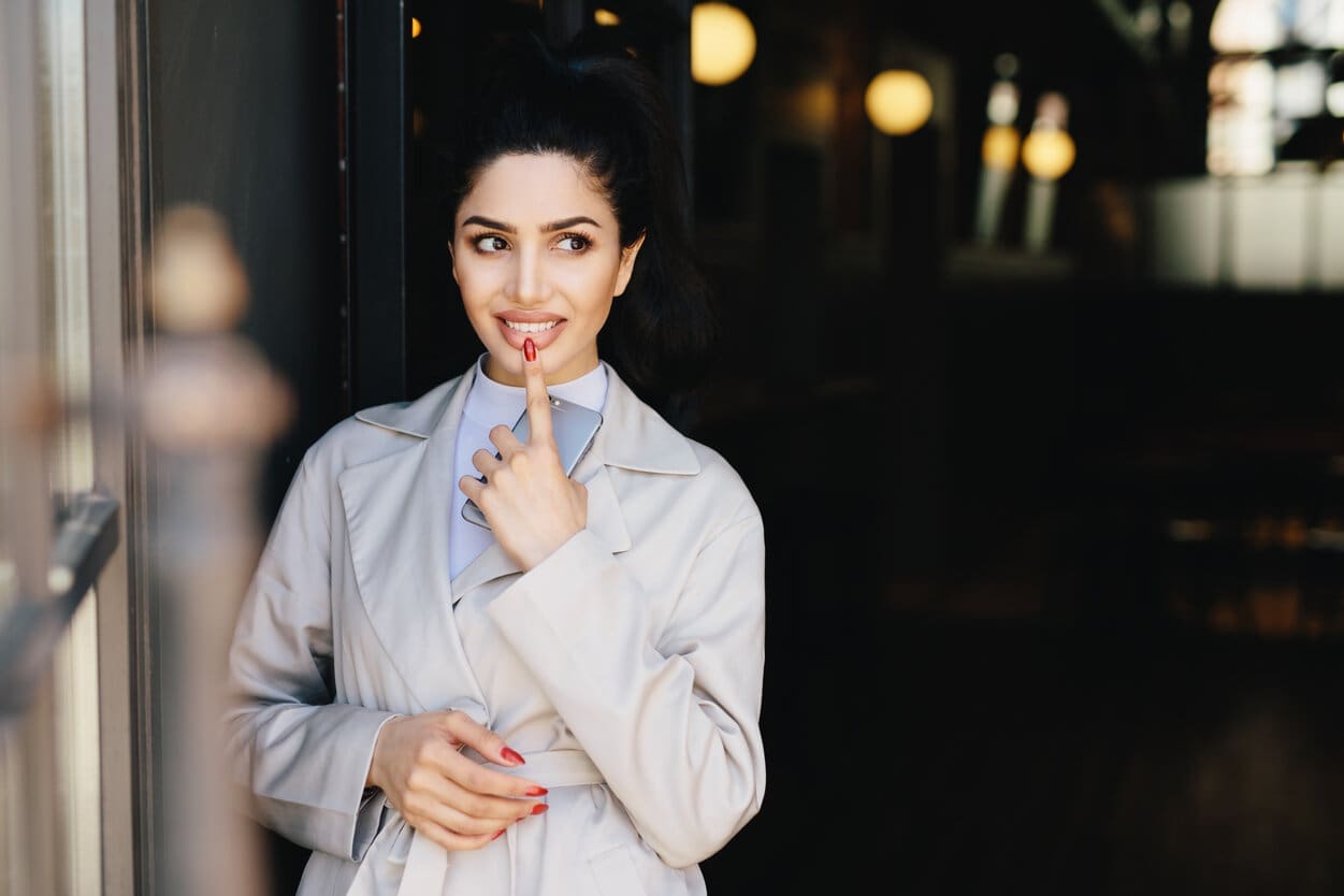 Fashionable lady with dark luminous hair tied in pony tail having nice eyes and red manicure holding her indexfinger on full lips and cell phone in hand having mysterious expression looking aside